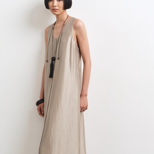 May include: A sleeveless, floor-length dress in a metallic champagne beige hue with a subtle sheen. The garment features a simple scoop neckline and a straight, flowing silhouette. A long black cord necklace with a tassel and a black bangle are worn.