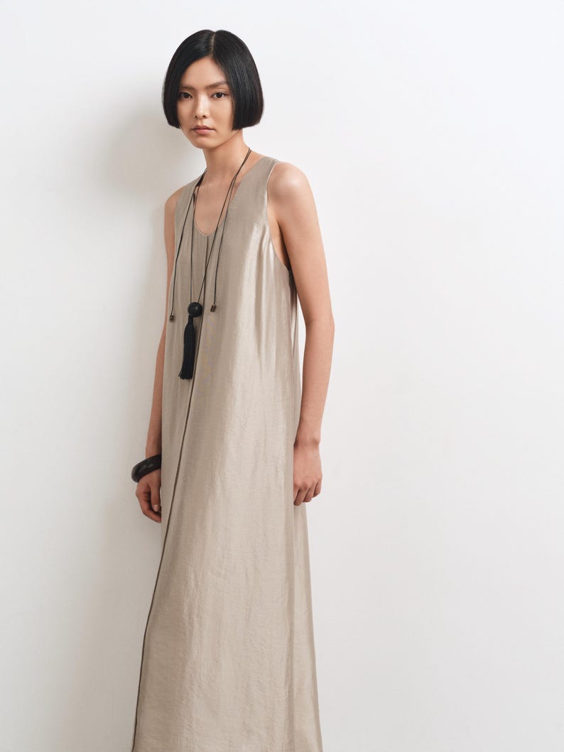 May include: A sleeveless, floor-length dress in a metallic champagne beige hue with a subtle sheen. The garment features a simple scoop neckline and a straight, flowing silhouette. A long black cord necklace with a tassel and a black bangle are worn.