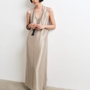 May include: A sleeveless, floor-length dress in a shimmering champagne-colored fabric with a scoop neckline. The garment is paired with a long, thin black cord necklace featuring a black tassel pendant and black mesh-style flat shoes.