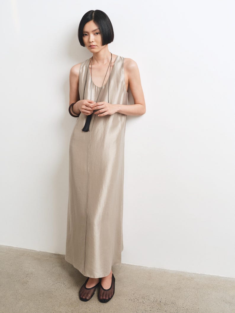 May include: A sleeveless, floor-length dress in a shimmering champagne-colored fabric with a scoop neckline. The garment is paired with a long, thin black cord necklace featuring a black tassel pendant and black mesh-style flat shoes.