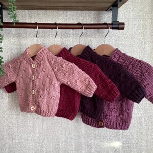 Puede incluir: Cinco cárdigans de bebé tejidos a mano en tonos rosa, rojo y morado cuelgan de perchas de madera. Los cárdigans tienen cierres de botón y patrones texturizados. Una planta decorativa cuelga de una estantería encima.