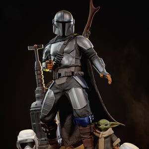 Puede incluir: Una estatua detallada de The Mandalorian, vestido con una armadura plateada, sosteniendo un bl&aacute;ster. La figura est&aacute; de pie con un rifle y una capa, acompa&ntilde;ada de una peque&ntilde;a figura de Grogu. La estatua est&aacute; sobre un fondo oscuro.