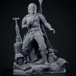 Puede incluir: Una figura gris de Mandaloriano con casco, armadura y capa, sosteniendo un arma. Una peque&ntilde;a figura de Grogu est&aacute; cerca. La base incluye un bl&aacute;ster, un casco y otros accesorios. La estatua es una representaci&oacute;n detallada.