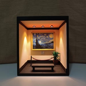 May include: Miniature diorama featuring a framed replica of Van Gogh's "Starry Night" painting. The scene includes a small bench, a potted plant, and a red velvet rope barrier, all within a lit, enclosed display.