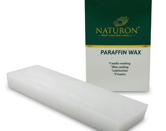 Op de afbeelding: Een witte blok paraffine was naast een groene en witte doos. Op de doos staat "NATURON" en "PARAFFIN WAX". De doos vermeldt ook de toepassingen van de was, waaronder kaarsen maken en het maken van kleurpotloden.