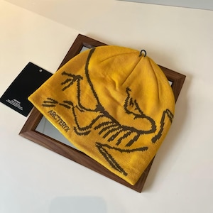 Może przedstawiać: Musztardowa czapka z brązowym wzorem graficznym i napisem "Arc'teryx" z przodu. Czapka spoczywa na lustrze w drewnianej ramie. Czarna metka jest widoczna w tle.