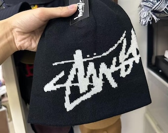 Puede incluir: Un gorro de punto negro con un gran logotipo de Stüssy blanco tejido en la tela. El gorro incluye una etiqueta de marca rectangular negra unida al borde superior con el logotipo de Stüssy impreso en texto blanco en el frente de la etiqueta.
