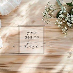 Könnte beinhalten: Ein heller Holzgrund mit einem weißen, durchsichtigen Stoff und einem Zweig weißer Blumen in der oberen rechten Ecke. Der Text "your design here" befindet sich in einem schwarzen Rechteck in der Mitte des Bildes.