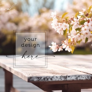 May include: A rustic wooden table with a whitewashed finish sits in a garden setting. A branch of delicate white blossoms hangs over the table, creating a natural and inviting scene. The text "your design here" is displayed in a transparent box on the table.