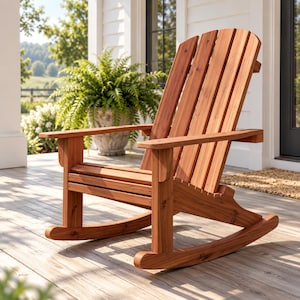 Op de afbeelding: Een houten schommelstoel met een hoge, latten rugleuning en brede armleuningen. De stoel is gemaakt van natuurlijk gekleurd hout met zichtbare nerven, voorzien van een gebogen zitting en twee gebogen schommelpoten aan de onderkant.