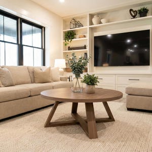 May include: A living room with a beige sofa, a round wooden coffee table, and a large flat-screen TV. The room has a neutral color palette with a white rug and built-in shelves. Decorative plants and pillows add accents.