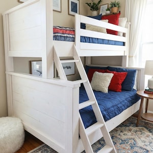May include: A white bunk bed with a ladder, featuring blue bedding, red and cream pillows. The room has framed artwork and a patterned rug. The bed frame has a slatted design.