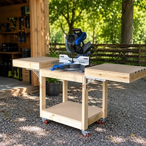 May include: A blue and black miter saw mounted on a light-colored wooden workbench with two folding side extensions. The workbench features a lower shelf and four red-wheeled casters for mobility, constructed from light-toned lumber.