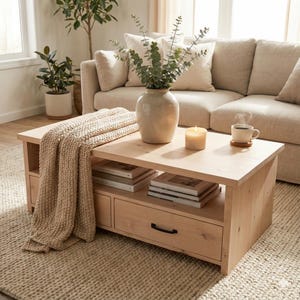 May include: A light wood coffee table with a lower shelf and drawer, holding books and magazines. A beige throw blanket is draped over the side. A vase with greenery, a lit candle, and a cup of coffee sit on top. A beige sofa is in the background.