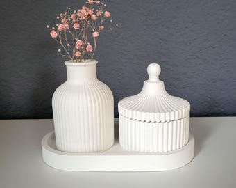 Könnte beinhalten: Ein weißes Keramik-Dekorationsset mit einer Vase mit rosa Blüten und einem Deckelglas, beide mit vertikalen Rippendetails. Die Artikel stehen auf einem passenden ovalen Tablett und bilden ein minimalistisches Ensemble.