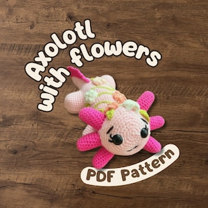 May include: A light pink crocheted axolotl plush toy featuring bright pink external gills, black button eyes, and small pastel-colored crocheted flowers attached to its back. The text Axolotl with flowers PDF Pattern is displayed.