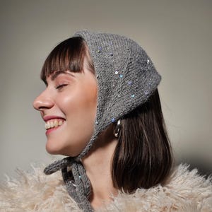 May include: A gray knitted head covering with sparkling sequins. The head covering is tied under the chin with a matching knitted tie. The person is wearing a cream-colored, fluffy coat and silver hoop earrings.