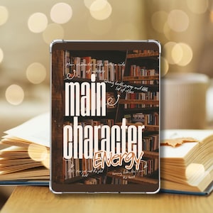 Puede incluir: Una funda para tableta con las palabras "Main Character Energy" en blanco. El fondo muestra una estantería llena de libros. La funda está sobre un libro abierto.
