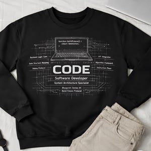 May include: Black sweatshirt with white text reading "CODE Software Developer System Architecture Specialist". The image includes khaki trousers, white trainers, and a laptop computer.