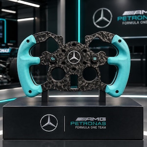 Puede incluir: Un volante de Fórmula 1 con marco de fibra de carbono negro y empuñaduras laterales de color turquesa brillante. El centro muestra un logotipo de Mercedes, con botones adicionales y una base negra con el logotipo de Mercedes-AMG Petronas Formula One Team.