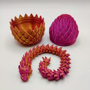 Puede incluir: Colección de artículos de fantasía impresos en 3D. Incluye un cuenco puntiagudo dorado y naranja, un recipiente en forma de huevo rosa y morado, y un dragón segmentado en colores a juego. Los objetos están sobre una superficie blanca.