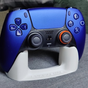 May include: A blue and black games controller with orange detailing, on a white stand. The controller has a textured finish and features various buttons and joysticks. The stand has the text "DUAL SENSE" on it.