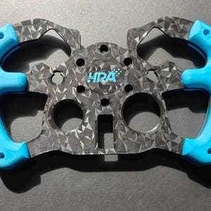 May include: A racing steering wheel rim featuring a black and gray geometric pattern with bright blue ergonomic side grips. The center of the wheel displays the text HRA in blue, with multiple circular mounting holes and cutouts throughout the frame.