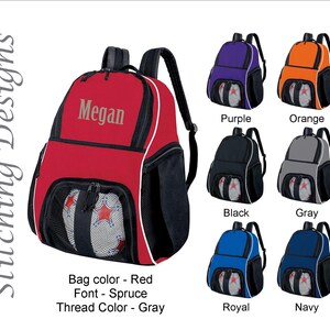 May include: A red backpack with the name "Megan" in gray. The bag has black mesh pockets and a white trim. Additional backpacks in purple, orange, black, gray, royal blue, and navy are also shown.