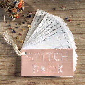 Puede incluir: Un libro de costura con una cubierta rosa y texto blanco. La cubierta dice "STITCH BOOK" con ilustraciones decorativas de puntadas. Las páginas están desplegadas, numeradas y sujetas con una cinta color crema. Flores secas están esparcidas.