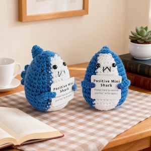 Peut inclure: Deux petits requins crochetés bleus et blancs, chacun tenant une pancarte avec l'inscription "Positive Mini Shark". Les pancartes indiquent "Always here to remind you: You're fin-tastic!". Ils sont posés sur un tissu à carreaux.