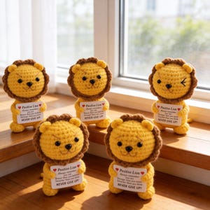 May include: Five handmade crochet lion figurines with brown manes and yellow bodies. Each lion holds a small sign that reads "Positive Lion" and a motivational message. The lions are displayed near a window.