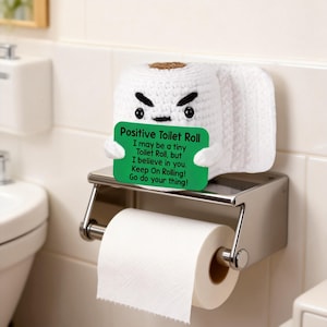 Op de afbeelding: Een witte gehaakte toiletrol met een boos gezicht, met een groen bord met de tekst "Positive Toilet Roll". Het bord heeft motiverende tekst. De rol staat op een chromen toiletpapierhouder.