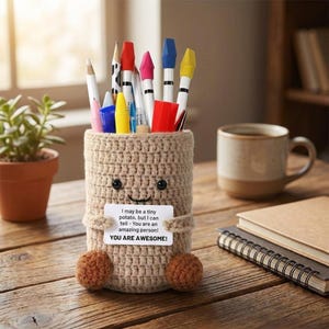Puede incluir: Un portalápices de ganchillo beige con forma de patata, con una cara sonriente y dos pequeños pies de bola marrones. El portalápices contiene varios bolígrafos y lápices. Una pequeña tarjeta dice: "I may be a tiny potato, but I can tell - You are an amazing person! YOU ARE AWESOME!"