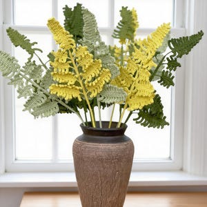 May include: A bouquet of handmade crochet fern stems in shades of green and yellow, arranged in a brown textured vase. The fern stems have detailed leaf patterns, creating a natural and decorative display. The vase is placed in front of a window.