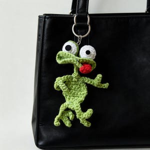 May include: A handmade green crochet frog keychain with white eyes and a red tongue. The frog is attached to a silver keyring, hanging against a black leather bag. The frog's arms and legs are outstretched.