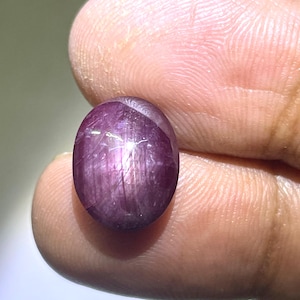 Può includere: Una gemma a cabochon di forma ovale con una tonalità rosso-viola intenso. La superficie mostra un distinto effetto a stella a sei raggi, noto come asterismo, visibile sulla cupola lucida e traslucida di questa pietra naturale.