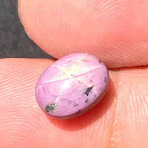 May include: A close-up of a polished, oval-shaped, pink gemstone. The stone has a star-like pattern across its surface and small dark inclusions. The gemstone is held between two fingers.