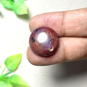 May include: A round, cabochon-cut ruby gemstone with a deep reddish-purple hue. The stone features a smooth, polished surface with visible natural inclusions, including small dark spots and a tiny surface indentation on the right side.