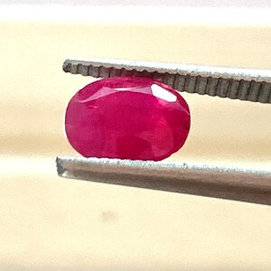 May include: A faceted, oval ruby gemstone held by tweezers. The ruby is a vibrant, deep red colour, with multiple facets catching the light. The tweezers are silver-coloured, and the background is a light beige.