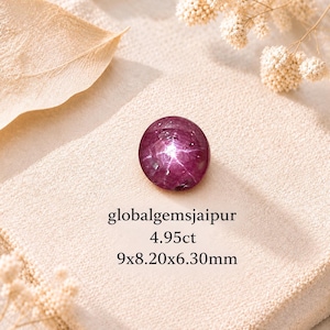 May include: A round, deep magenta star ruby gemstone featuring a visible six-rayed star effect. The text globalgemsjaipur, 4.95ct, and 9x8.20x6.30mm is printed below the stone. The gemstone displays a smooth, polished, cabochon-cut surface.