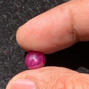 Peut inclure: Une petite pierre précieuse ronde, rose foncé, tenue entre deux doigts. La gemme a un aspect poli et translucide avec des inclusions internes en forme d'étoile. Le fond est une surface sombre et texturée.