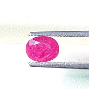 Può includere: Un rubino di forma ovale, tenuto da pinzette d'argento. Il rubino mostra un colore rosa intenso e saturo con motivi interni. La gemma è il punto focale, mostrando la sua superficie lucida e le caratteristiche naturali.