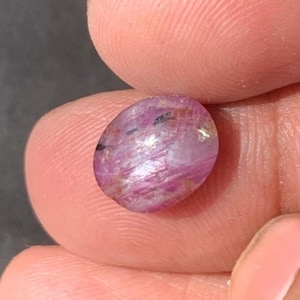 May include: A polished, oval-shaped gemstone with a pinkish-purple hue. The stone has internal veining and inclusions, with areas of lighter and darker color. The gem is held between two fingers, showcasing its size and detail.
