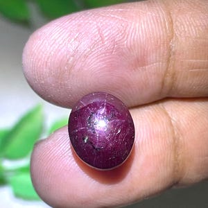 Könnte beinhalten: Ein tief magentafarbener Sternrubin-Edelstein mit einem glatten, polierten Cabochon-Schliff. Der Stein weist einen deutlichen sechsstrahligen Sterneffekt auf seiner Oberfläche auf, mit natürlichen Einschlüssen und einer durchscheinenden Qualität.