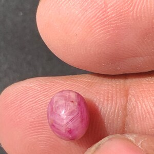 May include: A small, oval-shaped, pink gemstone with a translucent appearance. The gem has a polished surface with visible internal patterns and inclusions. The stone is held between two fingers, showcasing its size and color.