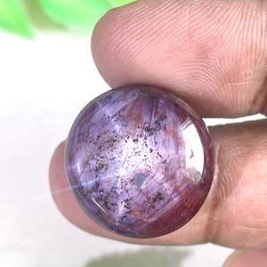 Peut inclure: Une pierre précieuse ronde taillée en cabochon présentant une palette de couleurs violet profond et rouge bordeaux. La surface affiche une texture fibreuse naturelle avec des inclusions sombres dispersées et une finition polie.