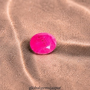 Könnte beinhalten: Ein facettierter, ovalförmiger, pinkfarbener Edelstein liegt auf einer strukturierten, hellbraunen Oberfläche. Die Abmessungen des Edelsteins betragen 4,7 x 5,85 x 3,3 mm. Der Text "globalgemsjaipur" ist enthalten.