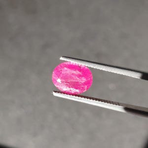 May include: A vibrant, oval-cut, hot pink gemstone is held by a pair of silver tweezers. The gemstone has a slightly textured surface and reflects light, showcasing its brilliant color. The background is a muted gray.