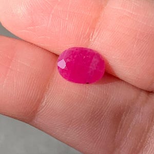 Peut inclure: Une pierre précieuse rose vif, taillée en forme ovale et à facettes. La gemme est tenue entre les doigts, mettant en valeur sa surface polie et sa couleur vibrante. Les facettes de la gemme captent la lumière, créant un effet scintillant.