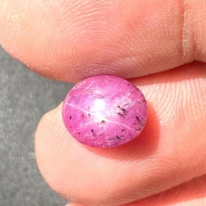 Può includere: Primo piano di una gemma rosa rotonda e lucidata. La gemma ha un effetto a stella con un centro luminoso e inclusioni scure. La gemma è tenuta tra due dita.